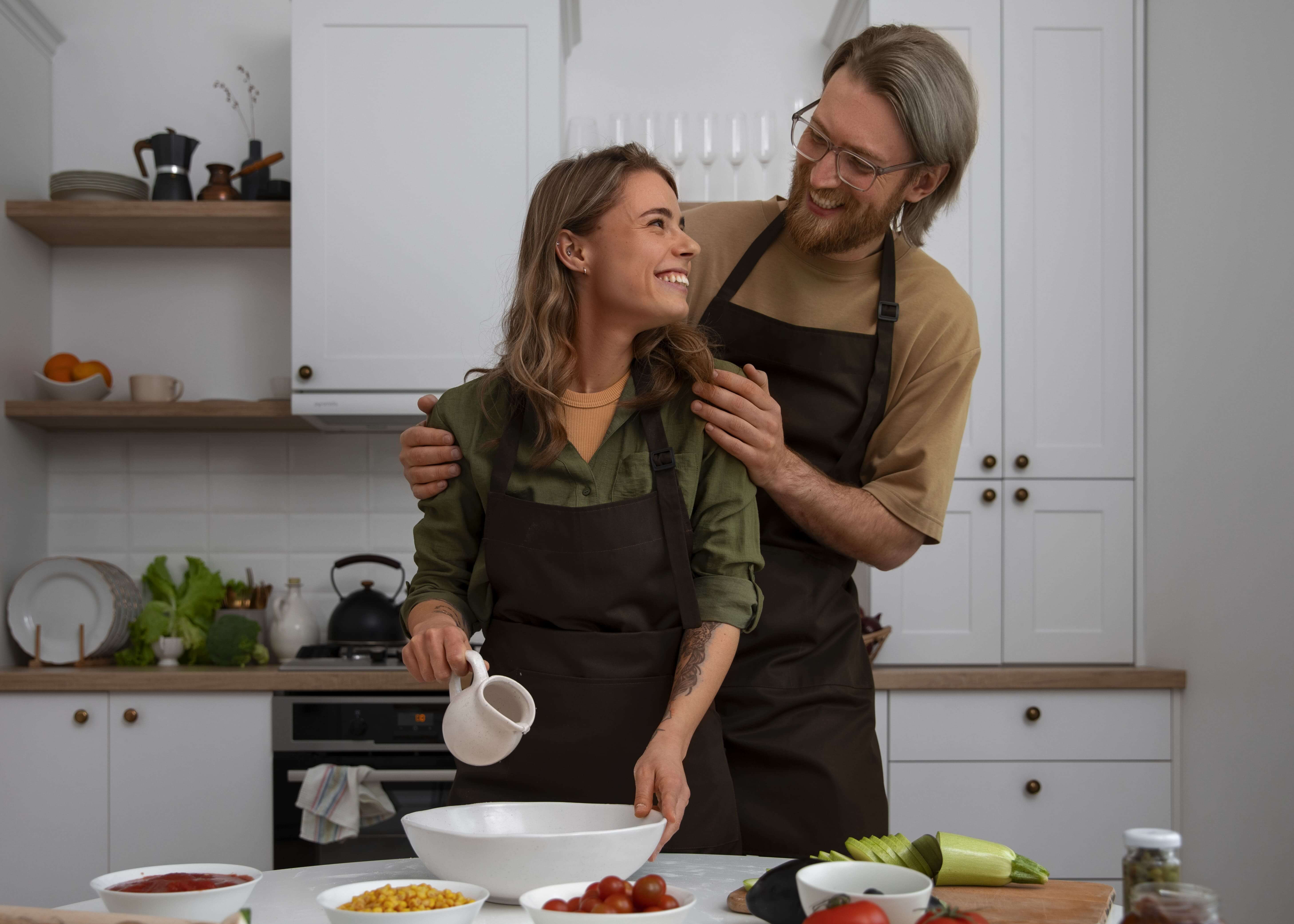 A man and woman wearing aprons stand together in a kitchen, engaged in cooking or preparing a meal.