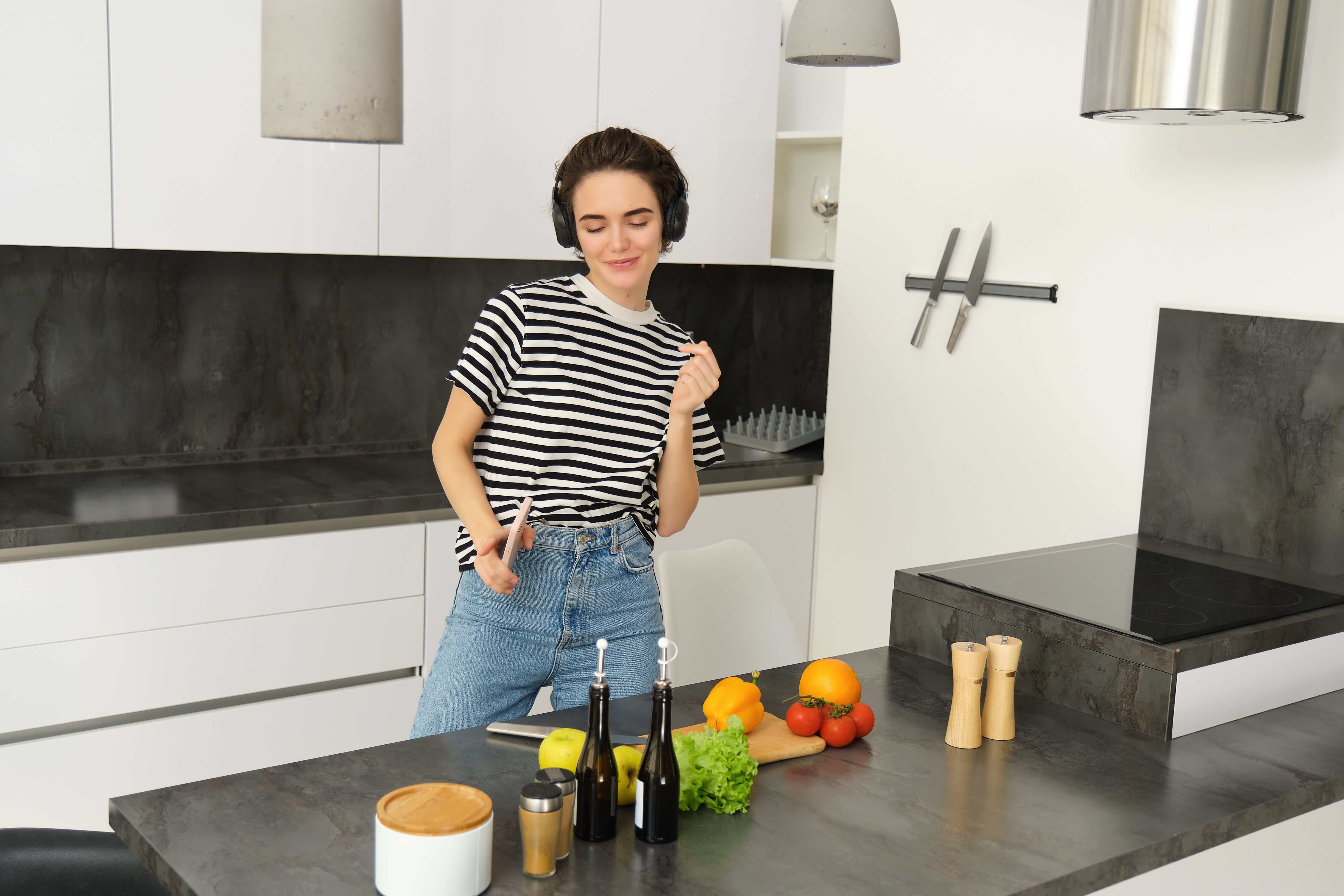 A woman in a kitchen holding a mobile phone, poised to begin her cooking process, surrounded by fresh ingredients.